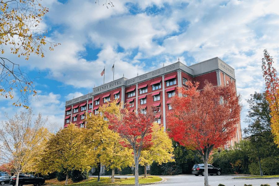 O.Henry Hotel facade surrounded by the contrasting colors of Autumn. Leaves of the trees are yellow, red, and orange. The photo promotes our new "Get Cozy Package" which includes: (1) Complimentary In-Room Movie (2) Indulgent Snack Pack — Choose Three: Granola Bars, Energy Bites, Cocoa Dusted Cashews, Cocoa Dusted Almonds, Buttered Popcorn, Truffle Popcorn, or Trail Mix (3) Warm Apple Cider for two.