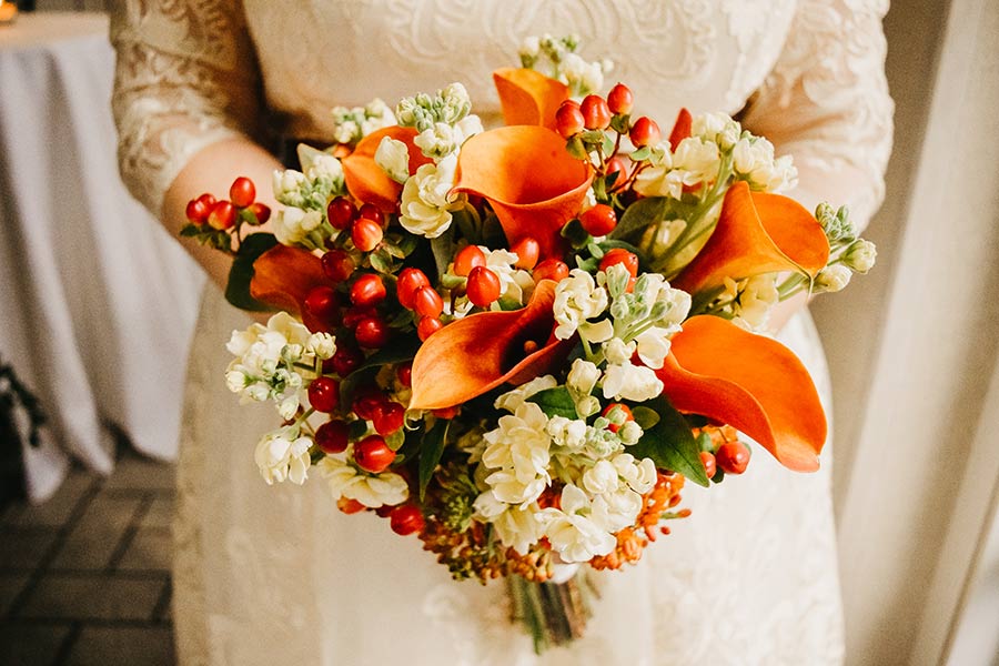 A bride holding a bouquet of peach, orange and white colored flowers with splashes of greenery.